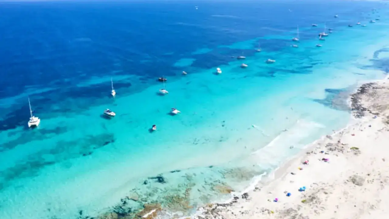 Aguas turquesas y arena blanca en una de las mejores playas de Formentera, con veleros y barcos anclados.