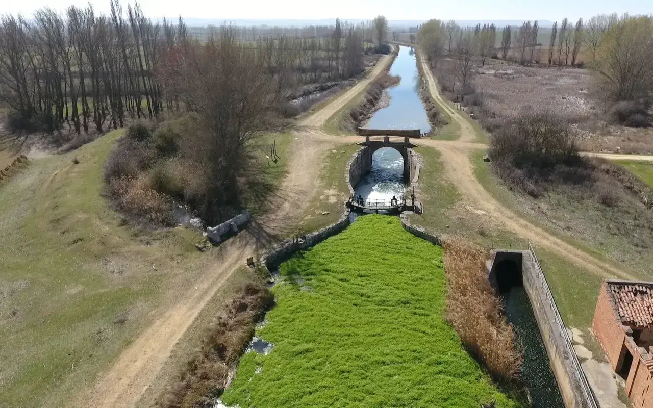 Canal de agua con vegetaci&oacute;n verde exuberante, un puente y caminos de tierra cerca de Boadilla del Camino.