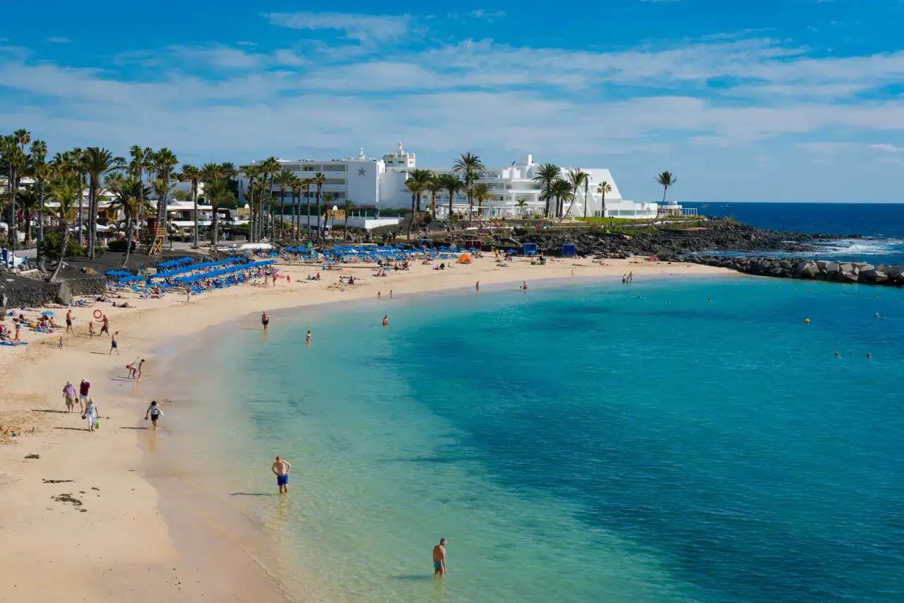 Una playa dorada con gente disfrutando del sol y el mar turquesa, rodeada de palmeras y hoteles blancos. Un paraíso entre las mejores playas de Lanzarote.