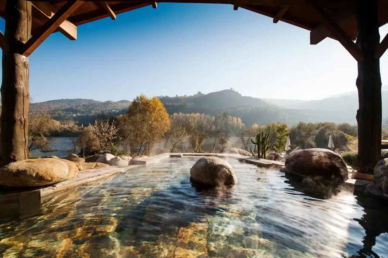 Rel&aacute;jate en estas termas en Galicia, con vapor ascendiendo del agua cristalina y un paisaje monta&ntilde;oso de fondo.