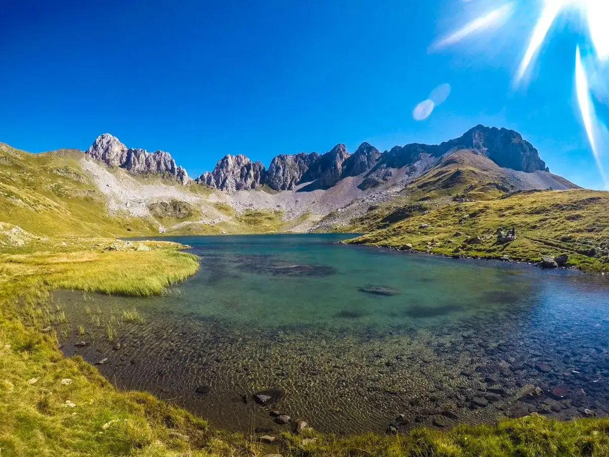 El ibón de Acherito, un lago de montaña de aguas cristalinas, rodeado de prados verdes y picos rocosos bajo un cielo azul intenso.