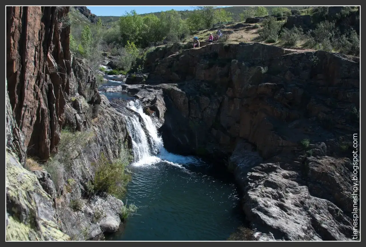 La cascada del aljibe desciende entre rocas y vegetación hacia una poza de aguas oscuras. Personas observan desde la cima.