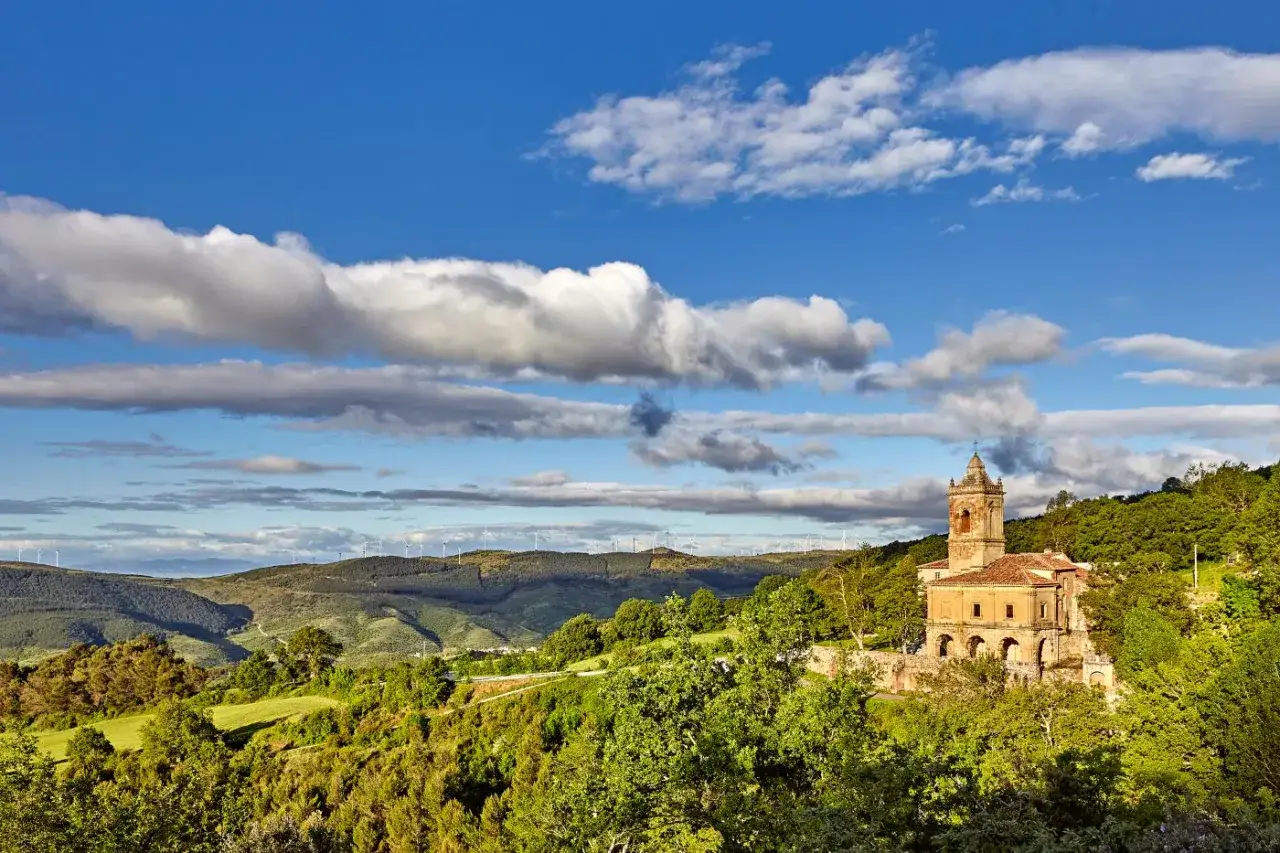Iglesia con torre campanario en una colina verde, rodeada de &aacute;rboles y con molinos de viento al fondo. Las torres del r&iacute;o vigilan el paisaje.