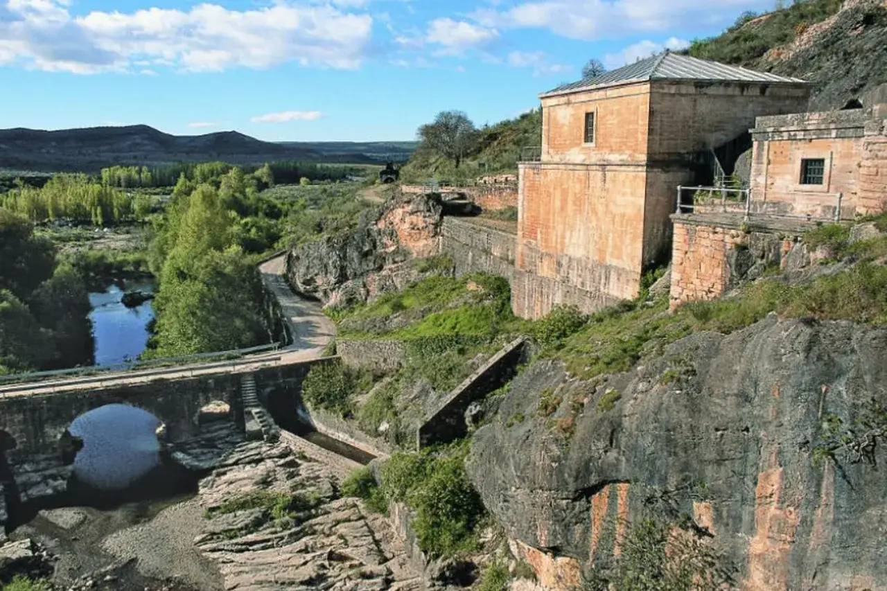 Antiguo pont&oacute;n de la oliva junto a un r&iacute;o y un puente de piedra, con edificios hist&oacute;ricos en la ladera rocosa.