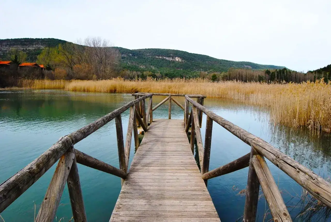 Muelle de madera que se adentra en la serena laguna de U&ntilde;a, rodeada de juncos dorados y colinas verdes.