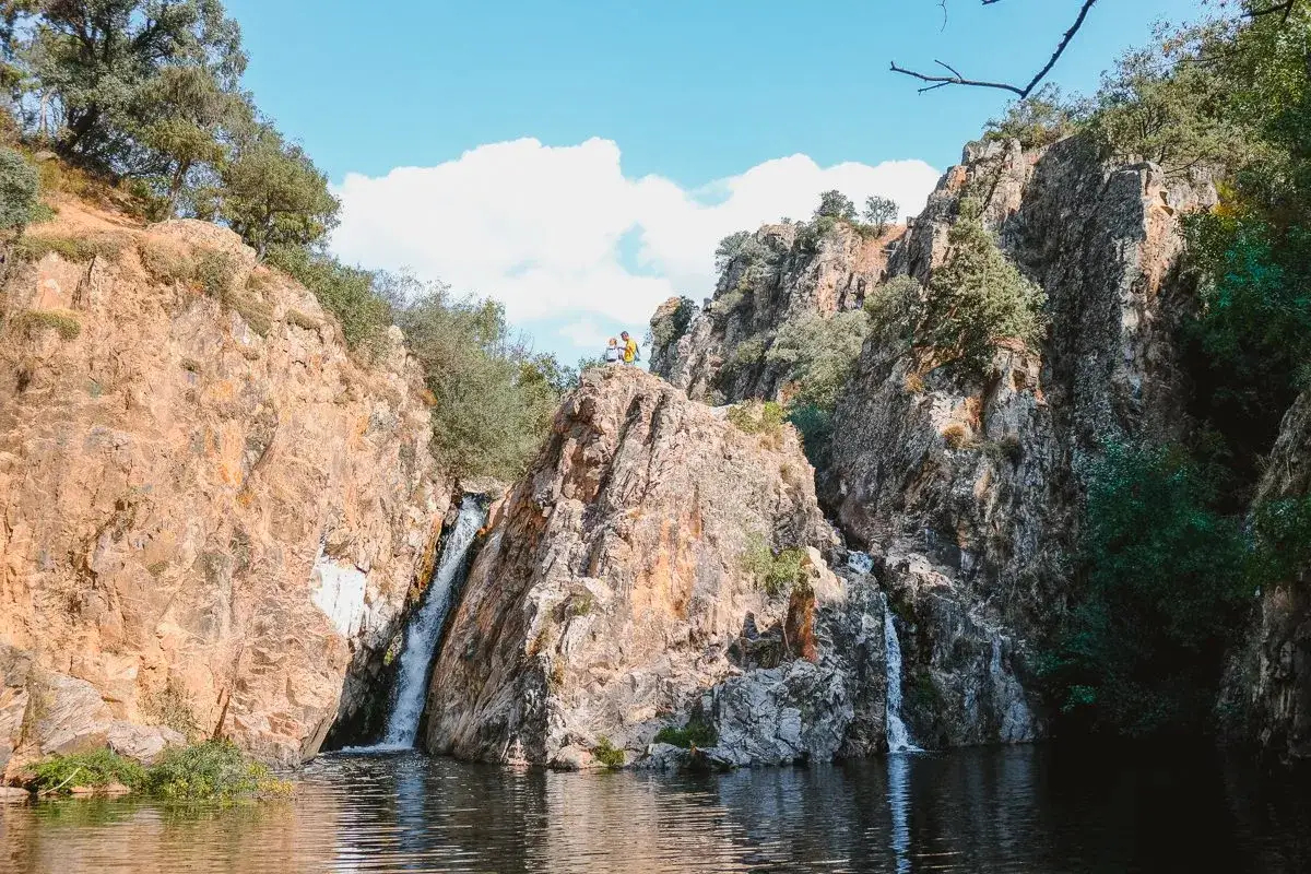 Dos personas admiran la cascada del hervidero, rodeada de rocas y vegetación exuberante bajo un cielo azul con nubes.