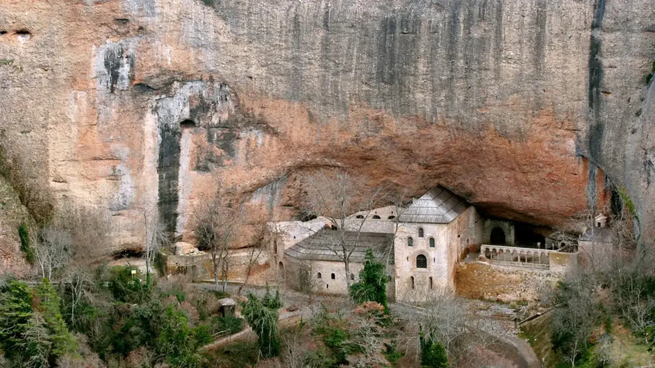 Monasterio de San Juan de la Peña, un impresionante conjunto arquitectónico incrustado en la roca, con sus edificios de piedra y tejados grises bajo un gran saliente rocoso.