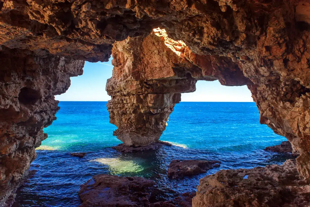Vista desde una cueva en Cala Moraig, con el mar azul intenso y rocas rojizas.