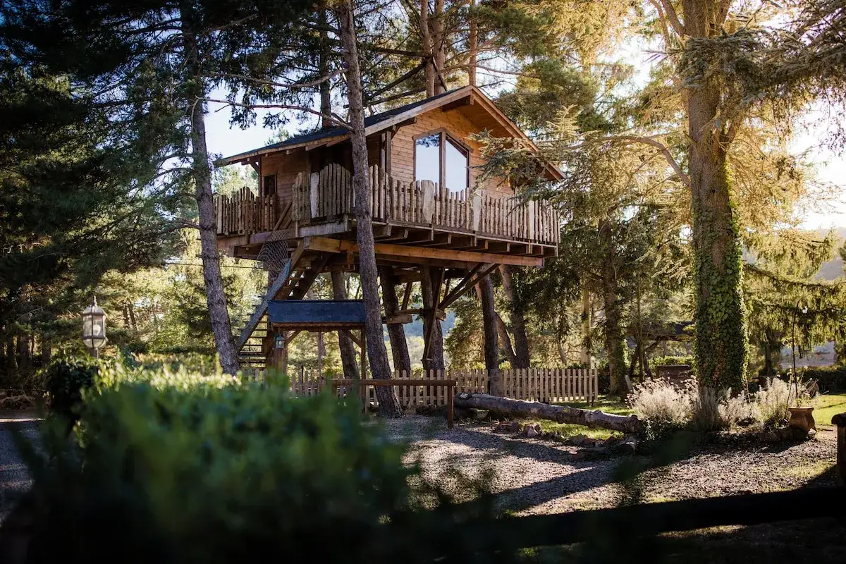 Una encantadora casa del árbol de madera, elevada entre árboles frondosos, con un balcón rústico y una escalera de acceso.