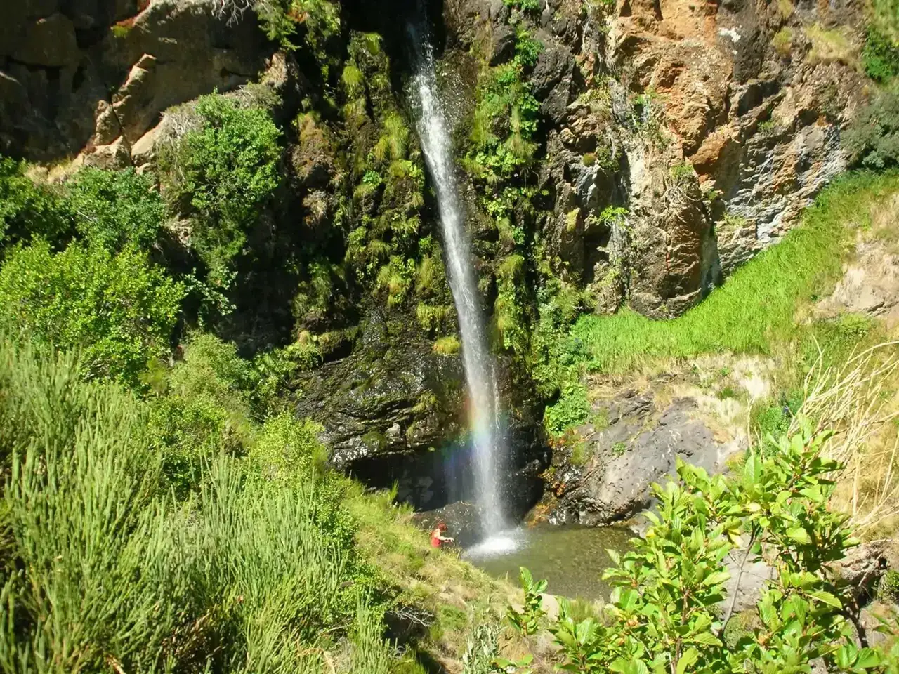 Una persona observa la cascada de Mazobre, con un arcoíris formado por el rocío. Vegetación exuberante rodea la caída de agua.