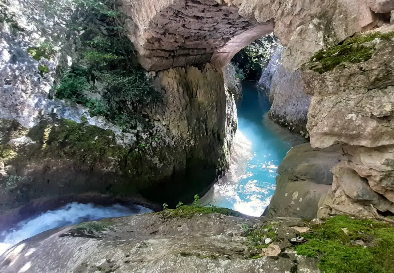 Agua turquesa fluye bajo un puente de piedra en el desfiladero del río Purón, rodeado de rocas cubiertas de musgo y vegetación.