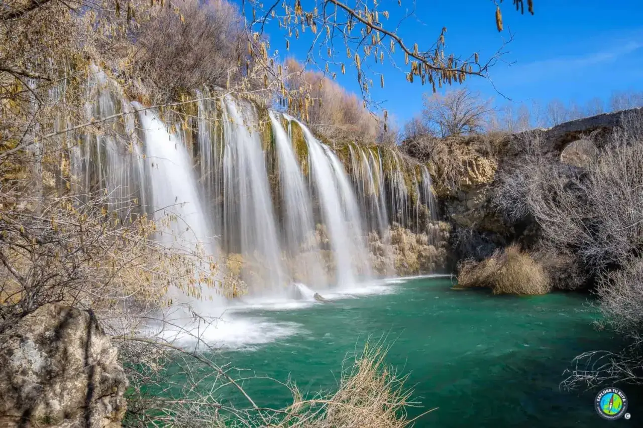 Cascada de agua turquesa, como los ojos del Cabriel, cayendo sobre rocas y rodeada de vegetación seca bajo un cielo azul.