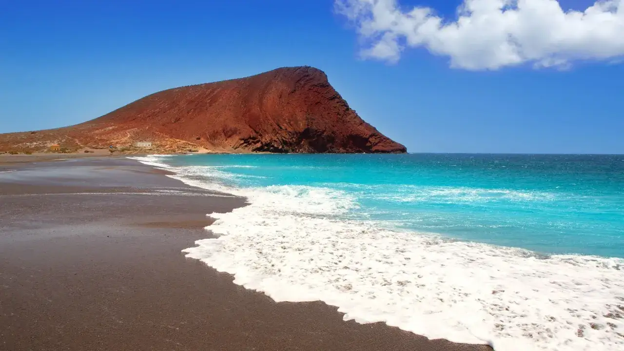 Playa de arena negra con el imponente cono volcánico de Montaña Roja al fondo. Aguas turquesas y espuma blanca, un paraíso entre las mejores playas de Tenerife.