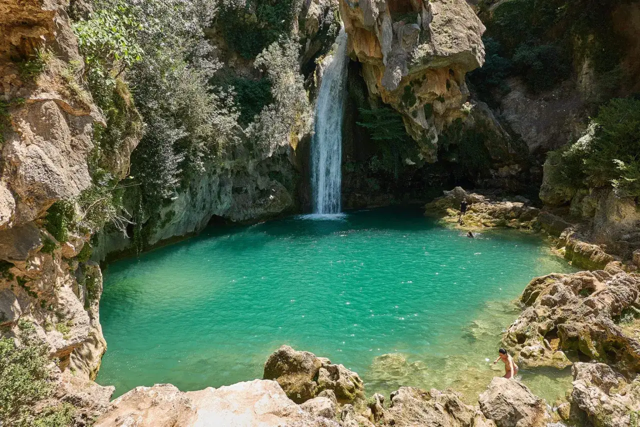 La cascada de la calavera desciende a una poza turquesa rodeada de rocas y vegetaci&oacute;n. Personas disfrutan del agua cristalina.