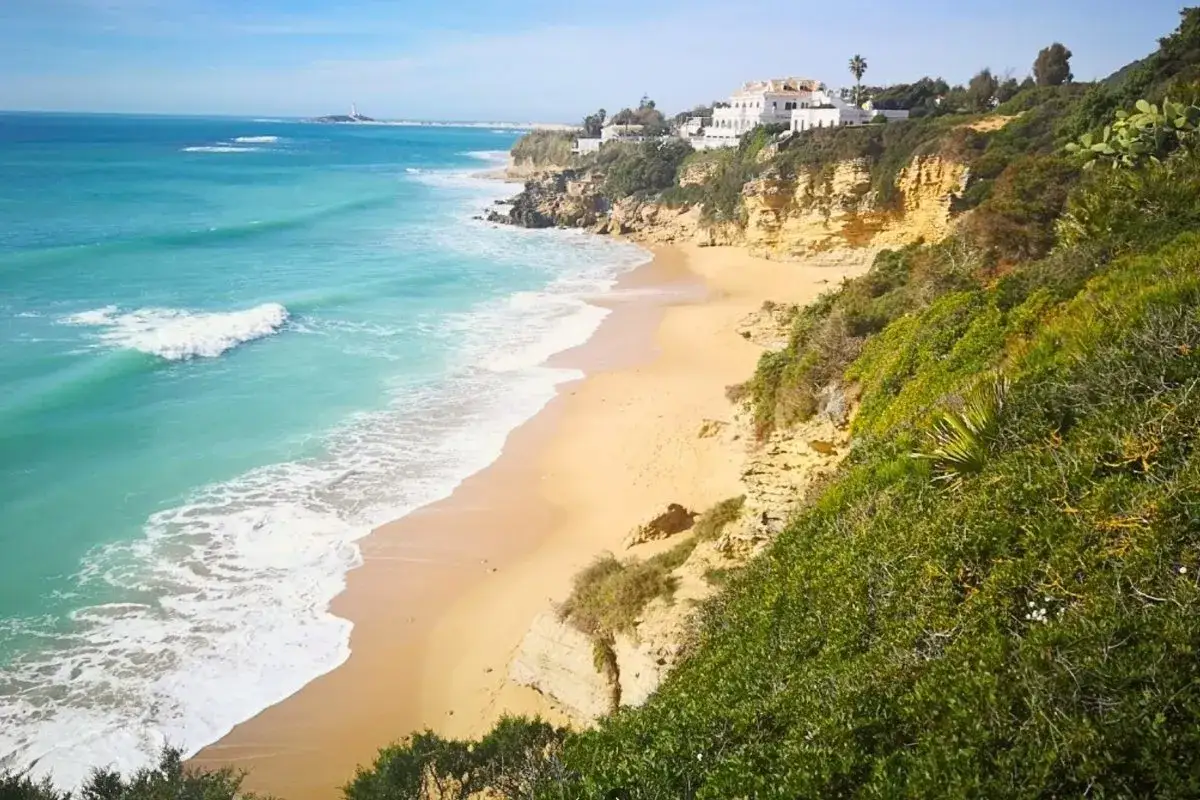 Mar turquesa y arena dorada en las playas de Cádiz, con acantilados verdes y casas blancas al fondo.