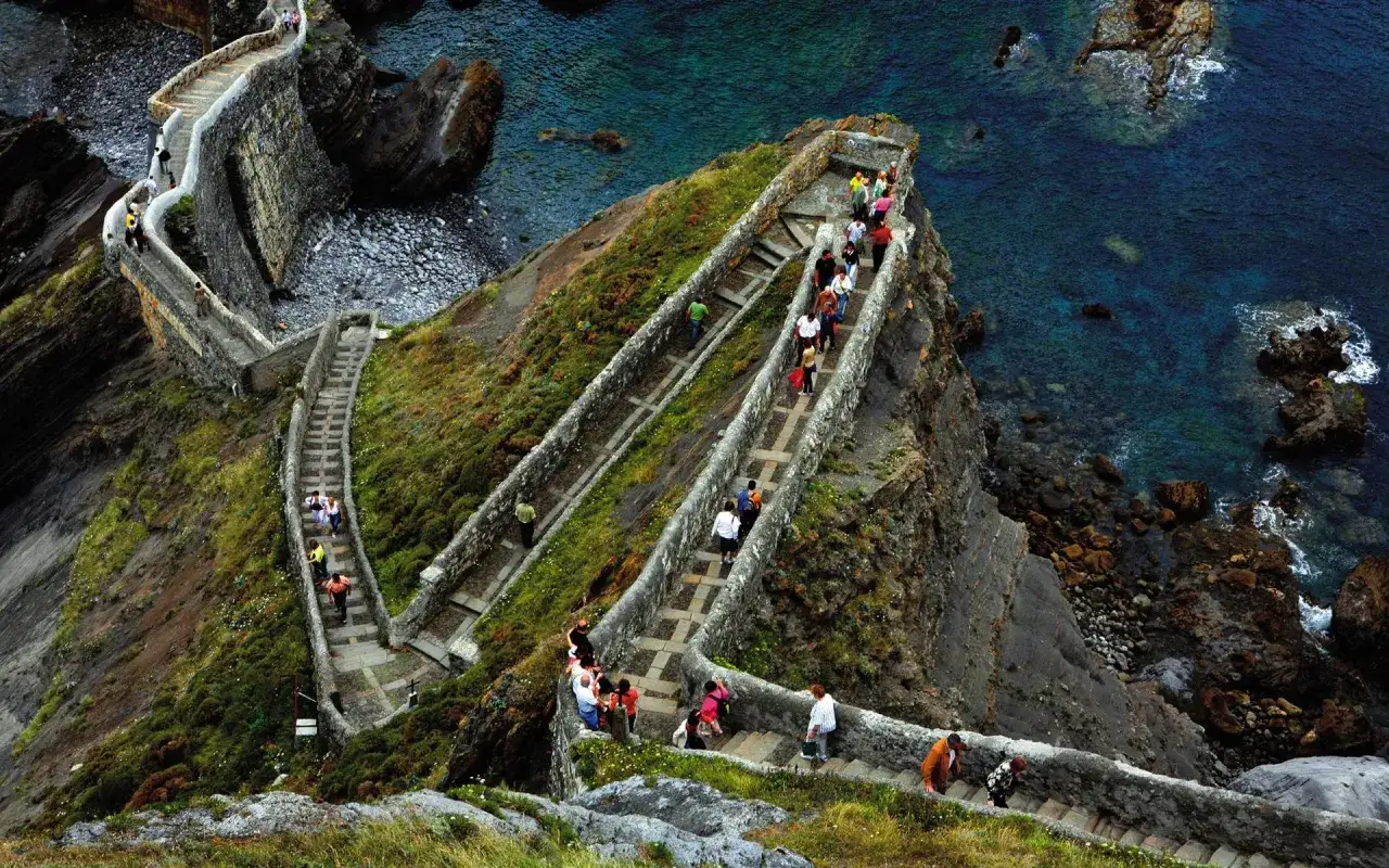 Ascenso por las empinadas escaleras de San Juan de Gaztelugatxe, con visitantes disfrutando del paisaje costero y el mar turquesa.