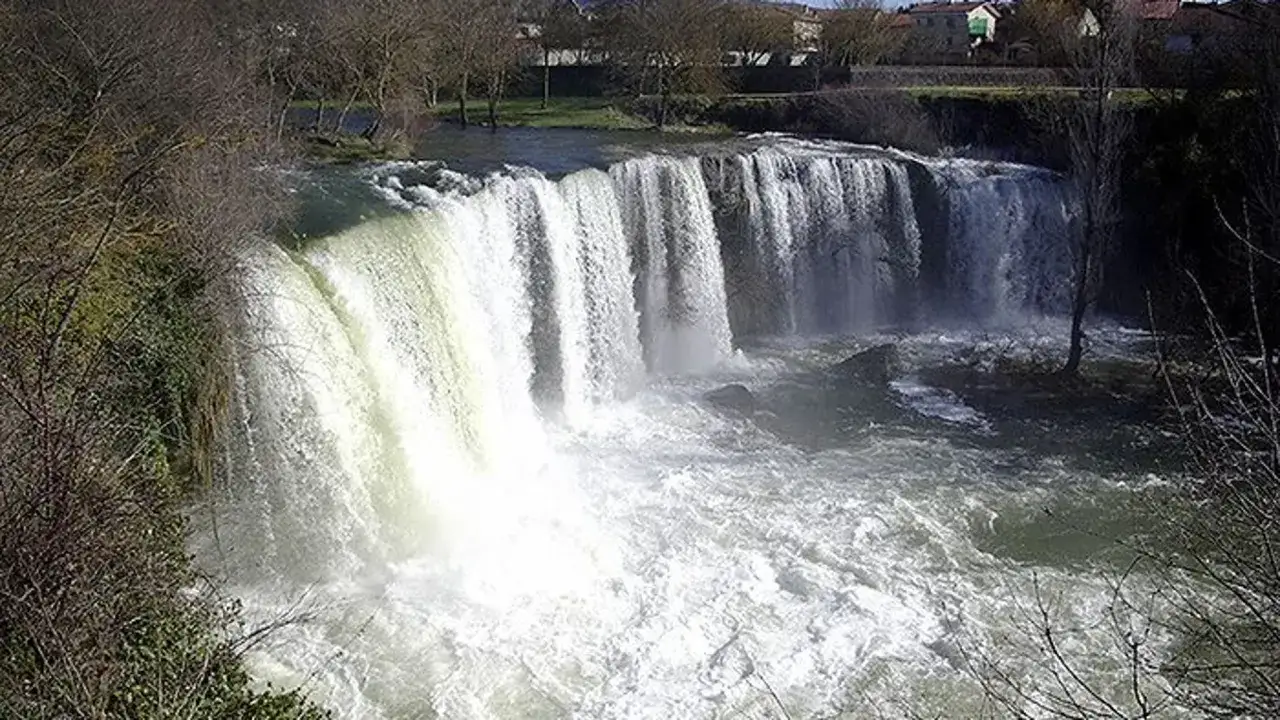 La imponente cascada de Tobalina, con su agua blanca y espumosa, cae sobre rocas y un río caudaloso. Al fondo, árboles desnudos y casas.