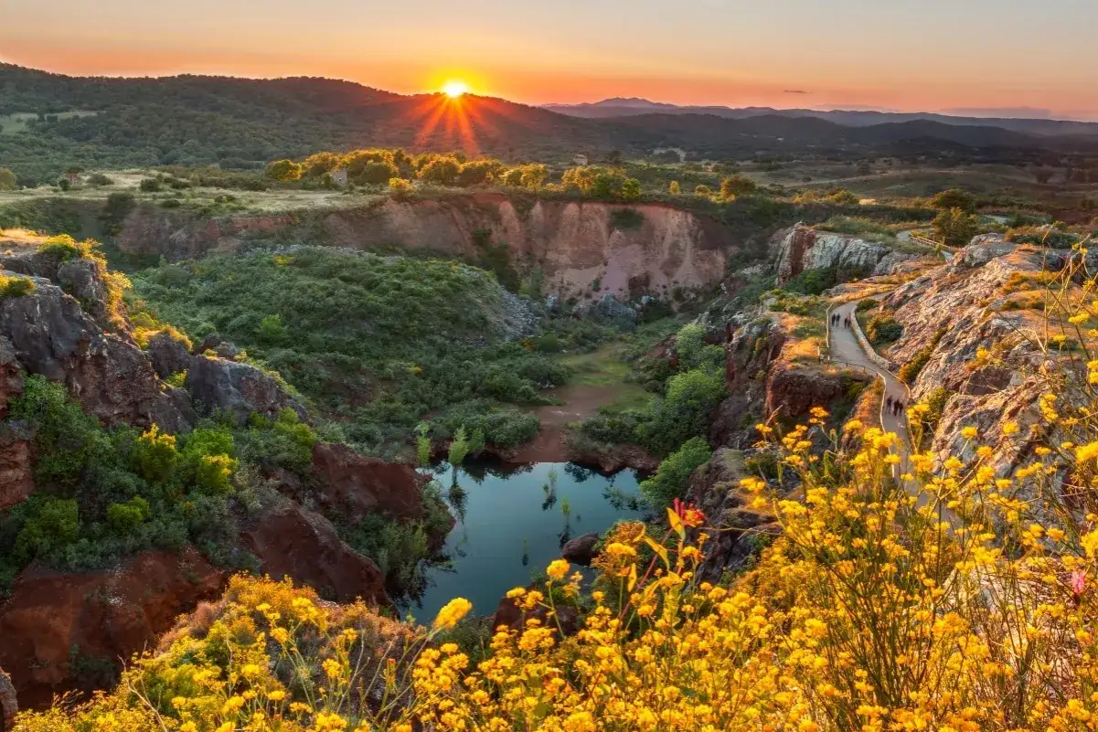 Atardecer en una ruta senderismo Sevilla. Un camino serpentea entre rocas y flores amarillas, con un lago en el fondo.