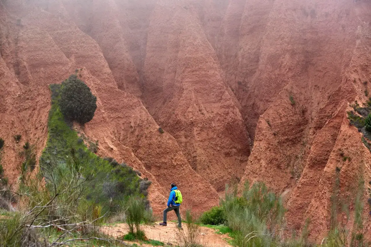 Senderista admira las imponentes carcavas Pont&oacute;n de la Oliva, un paisaje erosionado de tierra rojiza y vegetaci&oacute;n verde.