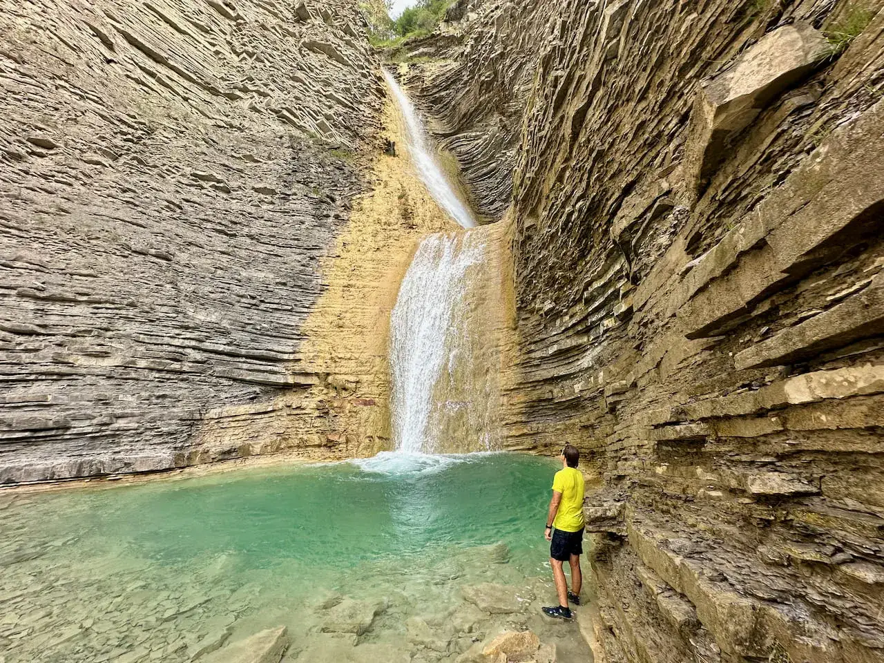 Hombre contempla una cascada de oros bajo, rodeada de paredes rocosas estratificadas y una poza de agua turquesa.