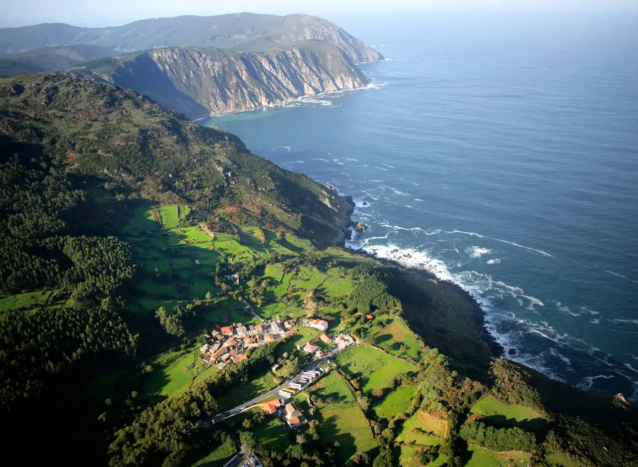 Vista aérea de San Andrés de Teixido, con sus acantilados imponentes y el mar azul. El pueblo se asienta entre verdes prados y bosques frondosos.