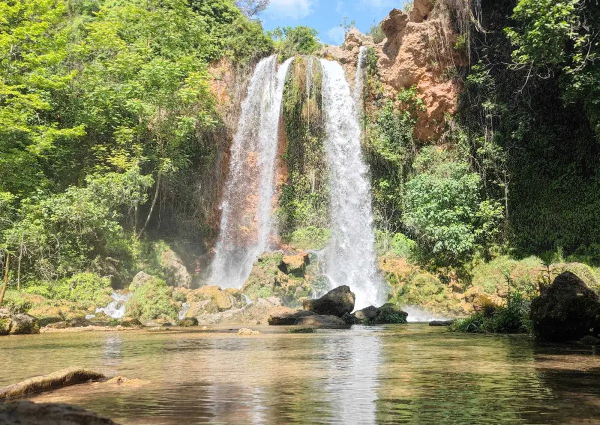 Cascada exuberante en los lagos de Anna, con agua cayendo sobre rocas y vegetaci&oacute;n verde. El sol ilumina el paisaje.