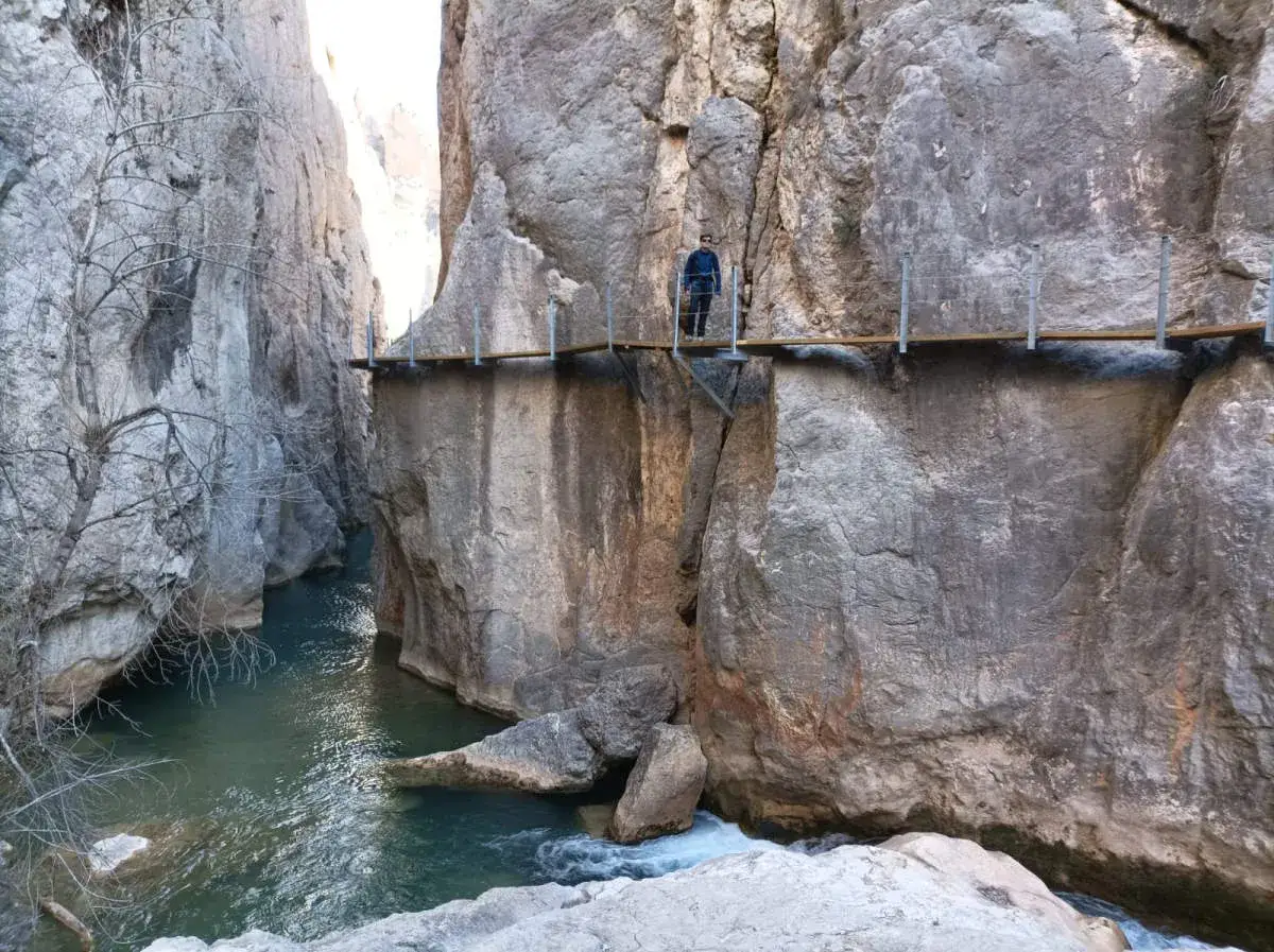 Un hombre camina por las pasarelas de Vallor&eacute;, un sendero suspendido sobre un r&iacute;o turquesa entre imponentes paredes rocosas.