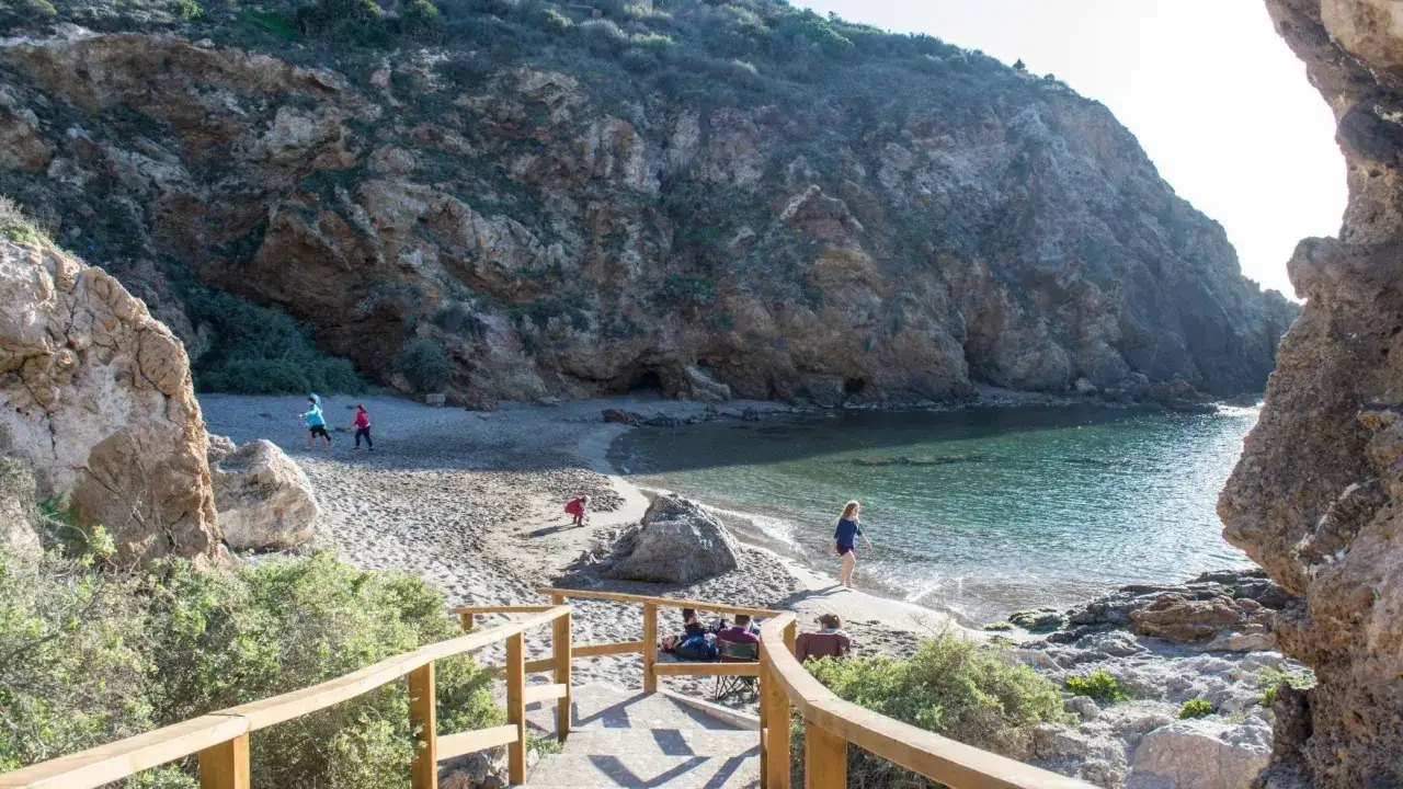 Escaleras de madera bajan a una cala rocosa, una de las mejores playas de Murcia. Niños juegan en la arena y el mar turquesa invita a un baño.