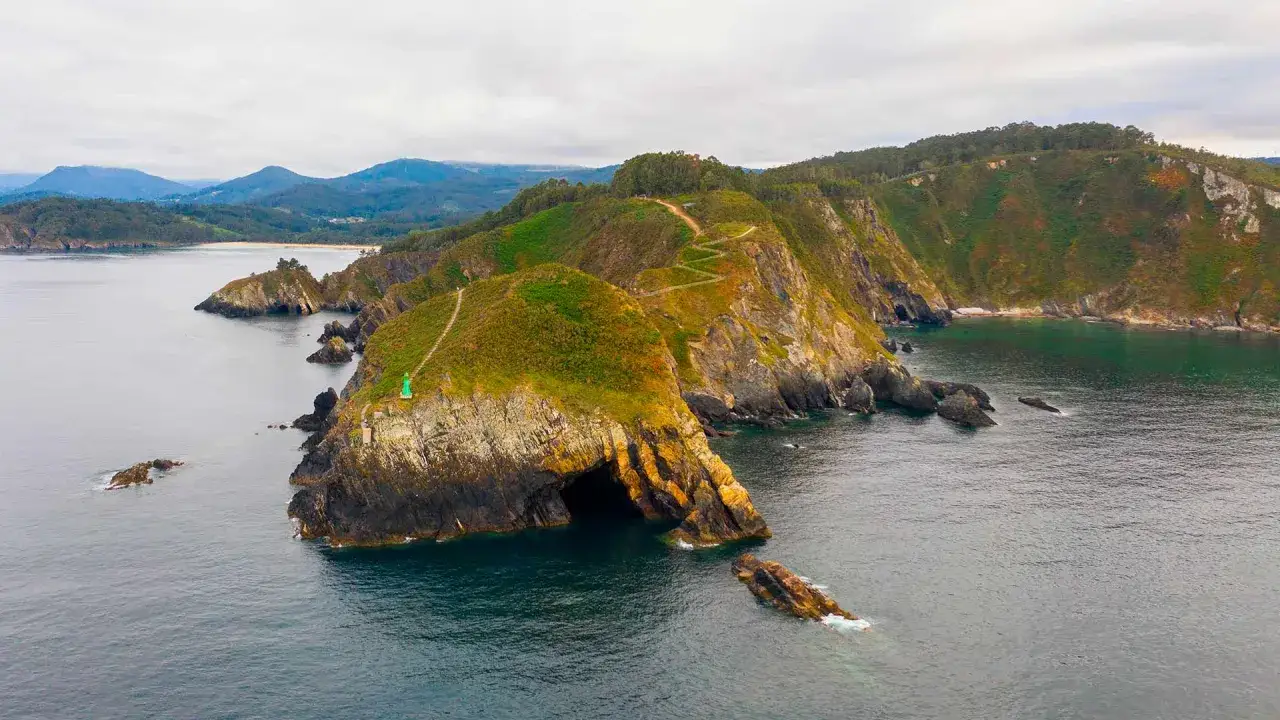 Costa rocosa con una cueva que parece el fuci&ntilde;o do porco. Senderos serpentean por las colinas verdes y un peque&ntilde;o faro verde vigila el mar.