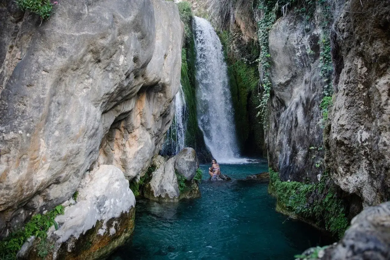 Una mujer se baña en las aguas turquesas de las fuentes del Algar, rodeada de cascadas y rocas cubiertas de vegetación.