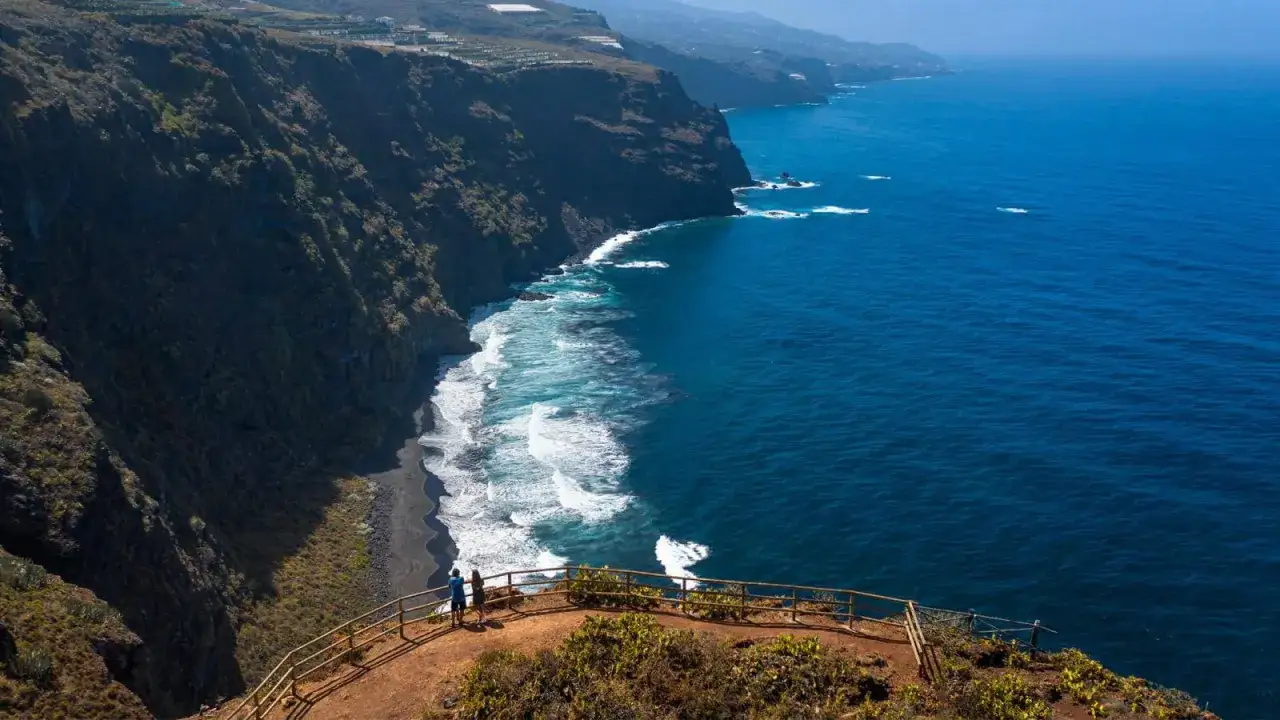 Dos personas admiran la playa de nogales, una costa de arena negra con olas rompiendo contra acantilados verdes.