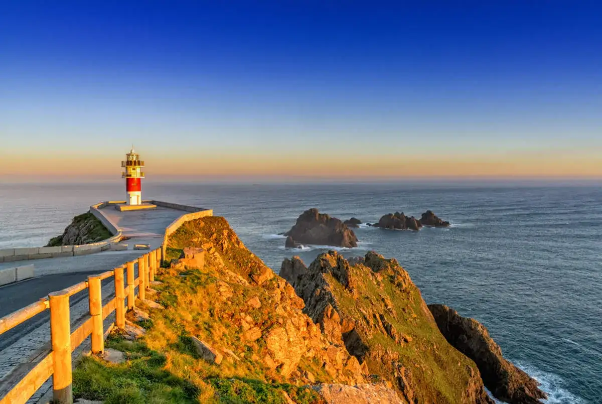 Faro rojo y blanco en un acantilado rocoso, con el mar azul y el cielo al atardecer. Un camino serpentea hacia él, ideal para una ruta por Galicia.