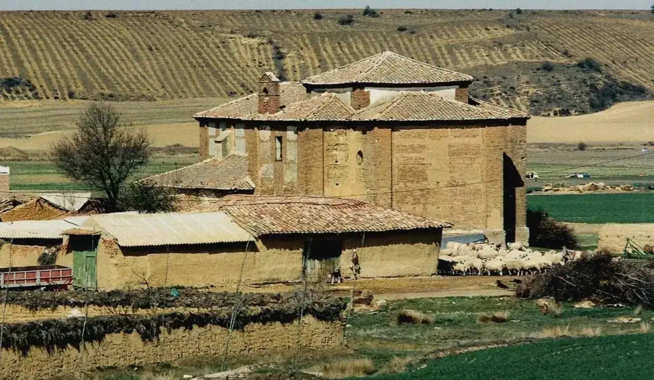 Edificio de ladrillo en Calzadilla de la Cueza, con reba&ntilde;o de ovejas pastando cerca. Campos labrados al fondo.