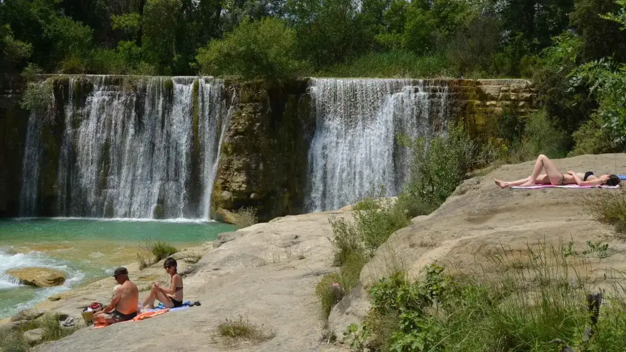 Pareja descansa junto a una cascada, mientras otra persona toma el sol en las rocas. Un día perfecto para disfrutar de la naturaleza y pozan de vero.
