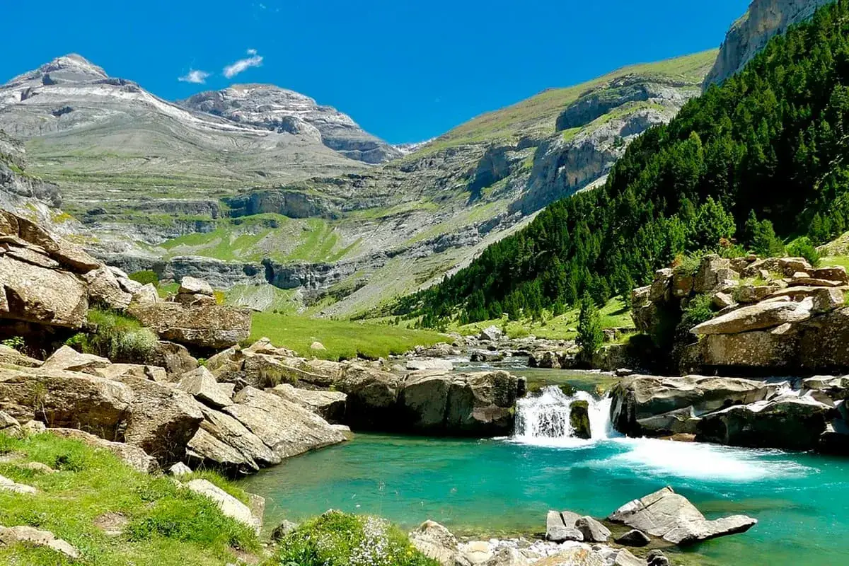 Un r&iacute;o turquesa serpentea entre rocas y prados verdes, con cascadas y monta&ntilde;as imponentes al fondo. Un paisaje id&iacute;lico que recuerda a los ibones de Anayet.