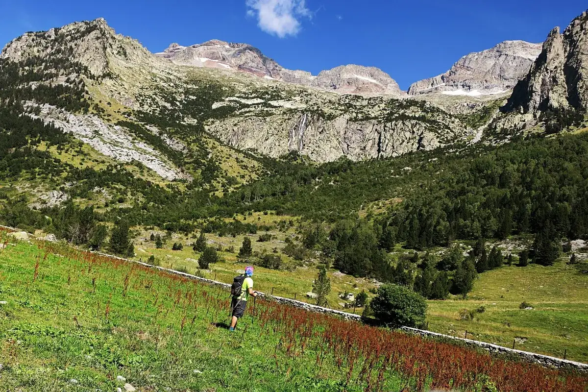 Senderista camina por un prado verde hacia imponentes monta&ntilde;as. A lo lejos, se vislumbra una cascada. El paisaje evoca la paz de una caba&ntilde;a de turmo.