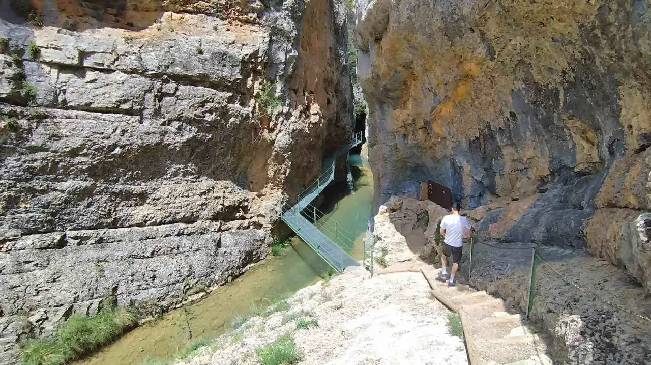 Un hombre camina por las pasarelas de Calomarde, un sendero metálico que serpentea sobre un río entre imponentes paredes rocosas.