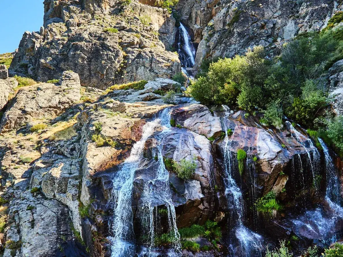 La cascada de los litueros desciende por rocas escarpadas, rodeada de vegetaci&oacute;n verde y bajo un cielo azul.