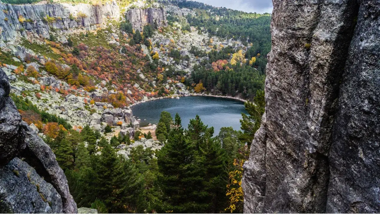 Paisaje otoñal con la laguna negra en el centro. Rocas y pinos rodean el lago, con colores rojizos y amarillos en la ladera. Para llegar, sigue el sendero entre las rocas.