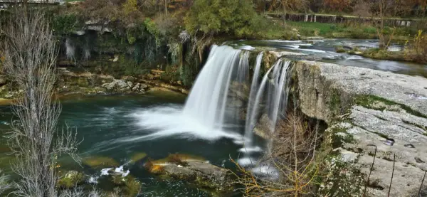 La majestuosa cascada de Tobalina, con su velo de agua cayendo sobre rocas cubiertas de musgo, rodeada de árboles otoñales y un río sereno.